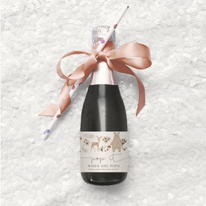 Puede incluir: Una peque&ntilde;a botella de champ&aacute;n con una cinta rosa y una pajita. La botella tiene una etiqueta que dice "pop it when she pops" y presenta una ilustraci&oacute;n de acuarela de un oso, un ciervo y flores.