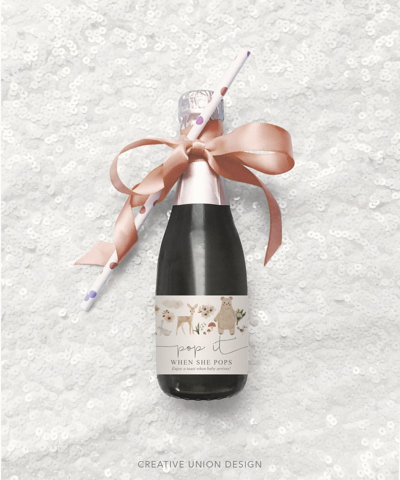 Puede incluir: Una peque&ntilde;a botella de champ&aacute;n con una cinta rosa y una pajita. La botella tiene una etiqueta que dice "pop it when she pops" y presenta una ilustraci&oacute;n de acuarela de un oso, un ciervo y flores.