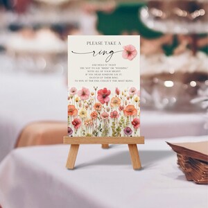 May include: A white card with a floral design and the text "PLEASE TAKE A ring AND HOLD IT TIGHT TRY NOT TO SAY "BRIDE" OR "WEDDING" WITH ALL OF YOUR MIGHTI IF YOU HEAR SOMEONE SAY IT. SNATCH UP THEIR RING TO WIN AT THE END. COLLECT THE MOST BLING." The card is on a small wooden easel.