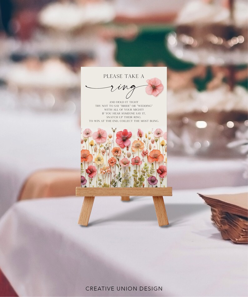May include: A white card with a floral design and the text "PLEASE TAKE A ring AND HOLD IT TIGHT TRY NOT TO SAY "BRIDE" OR "WEDDING" WITH ALL OF YOUR MIGHTI IF YOU HEAR SOMEONE SAY IT. SNATCH UP THEIR RING TO WIN AT THE END. COLLECT THE MOST BLING." The card is on a small wooden easel.