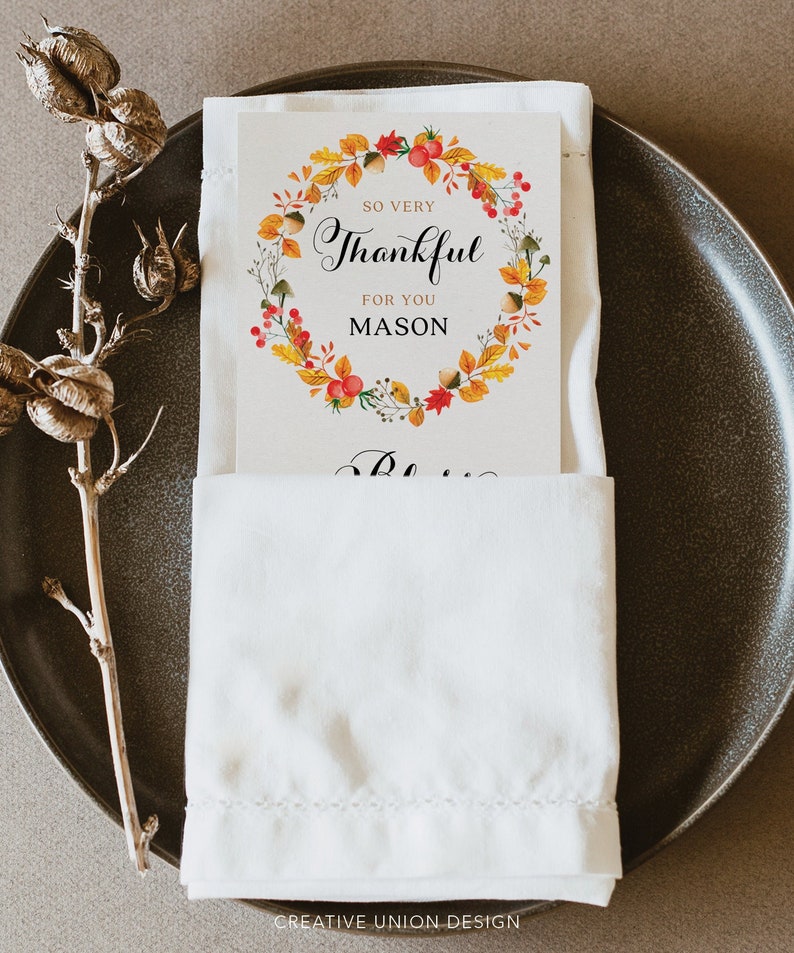 May include: A white napkin with a folded card on top. The card has a wreath of fall leaves and berries with the text "So very Thankful for you Mason".