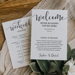 May include: Two white cards with black calligraphy text. One card says "Weekend Events" with a schedule. The other says "Welcome, We're so happy you're here" and includes a message for family and friends. Green leaves and a white flower are in the foreground.