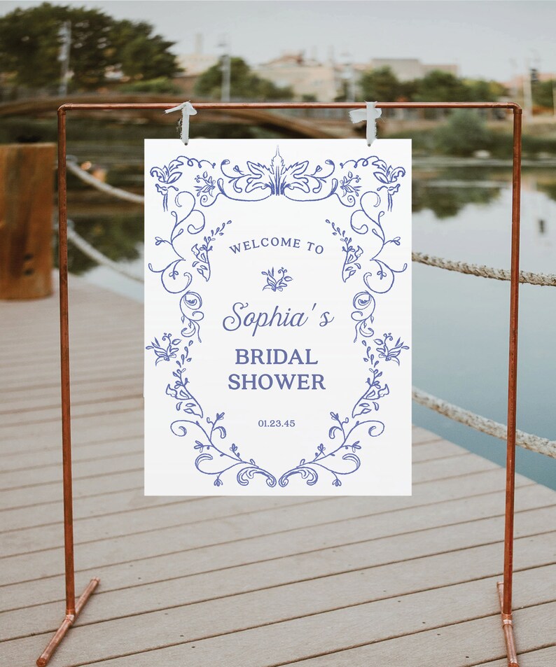 May include: White sign with a blue floral border and the text "Welcome to Sophia's Bridal Shower 01.23.45". The sign is hanging from a copper stand.