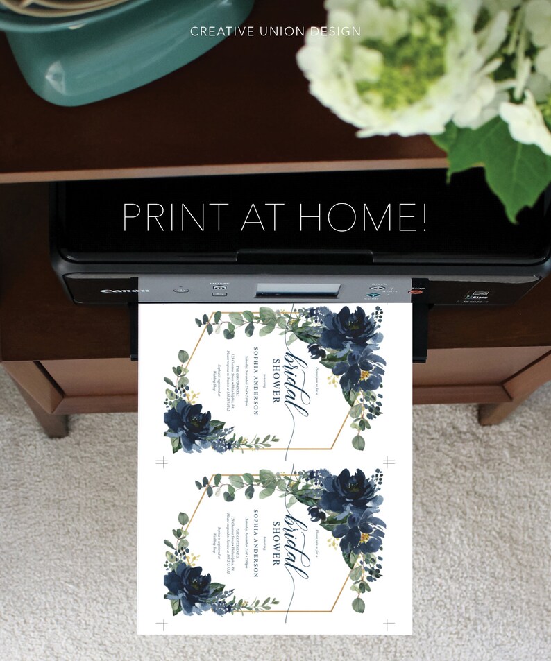 Op de afbeelding: Een printer drukt twee uitnodigingen voor een bruidsdouche af. De uitnodigingen hebben marineblauwe bloemstukken, gouden geometrische frames en de tekst "PRINT AT HOME!" en "bridal shower". Het ontwerp bevat de tekst "SOPHIA ANDERSON".