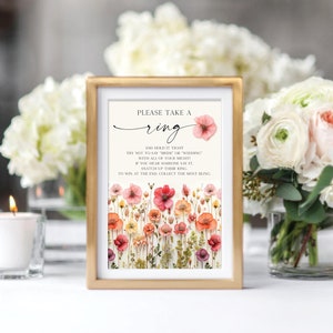 May include: A framed sign with the text "PLEASE TAKE A ring AND HOLD IT TIGHT TRY NOT TO SAY "BRIDE" OR "WEDDING" WITH ALL OF YOUR MIGHTY IF YOU HEAR SOMEONE SAY IT, SNATCH UP THEIR RING. TO WIN AT THE END, COLLECT THE MOST BLING."  The sign is decorated with pressed flowers.