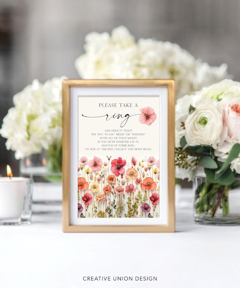 May include: A framed sign with the text "PLEASE TAKE A ring AND HOLD IT TIGHT TRY NOT TO SAY "BRIDE" OR "WEDDING" WITH ALL OF YOUR MIGHTY IF YOU HEAR SOMEONE SAY IT, SNATCH UP THEIR RING. TO WIN AT THE END, COLLECT THE MOST BLING."  The sign is decorated with pressed flowers.