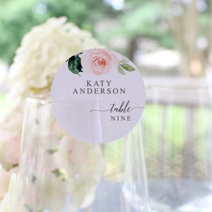 May include: A light purple round table number card with a watercolor floral design. The card says "Katy Anderson table nine".