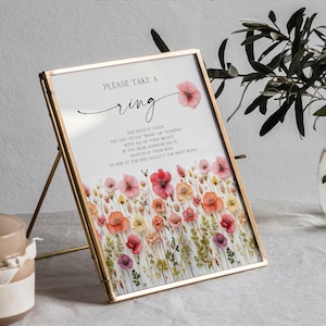 K&ouml;nnte beinhalten: Ein gerahmtes Schild mit einem goldenen Metallrahmen, das den Text "PLEASE TAKE A ring" und Anweisungen f&uuml;r ein Hochzeitsspiel enth&auml;lt. Das Schild ist mit Aquarellblumen in Rosa-, Rot- und Gelbt&ouml;nen verziert. Der Text "PRINTED & READY TO FRAME" steht oben.