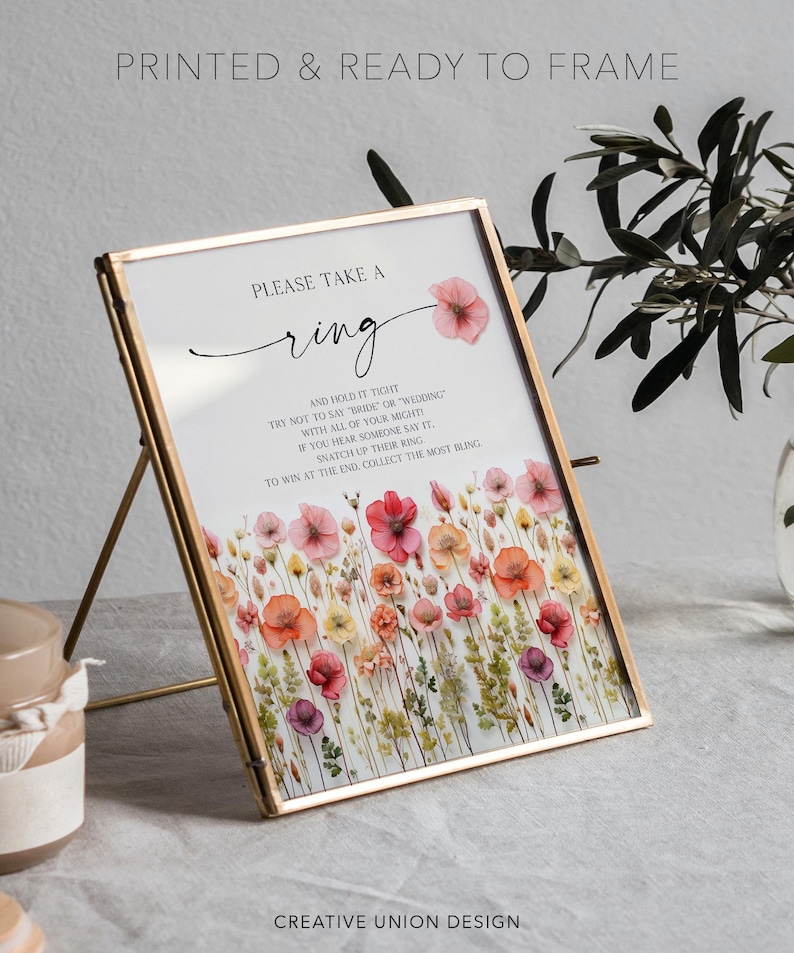 K&ouml;nnte beinhalten: Ein gerahmtes Schild mit einem goldenen Metallrahmen, das den Text "PLEASE TAKE A ring" und Anweisungen f&uuml;r ein Hochzeitsspiel enth&auml;lt. Das Schild ist mit Aquarellblumen in Rosa-, Rot- und Gelbt&ouml;nen verziert. Der Text "PRINTED & READY TO FRAME" steht oben.