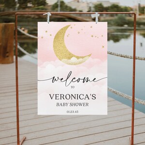 May include: A rectangular welcome sign featuring a gold glitter crescent moon and stars over pink watercolor clouds. The text reads: welcome TO VERONICA'S BABY SHOWER 01.23.45. The sign is suspended from a copper-colored metal frame stand.