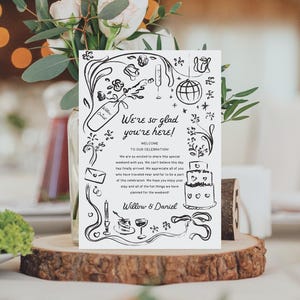 以下が含まれることがあります： 「We're so glad you're here! 」と「Willow & Daniel」のテキストが書かれた、白黒の結婚式用ウェルカムサインのイラスト。イラストには、シャンパングラス、ミラーボール、ケーキ、その他の結婚式に関連するイメージが含まれています。