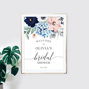 May include: A rectangular sign in a gold frame featuring a watercolor floral arrangement of navy, light blue, and peach flowers at the top. The text reads: WELCOME TO OLIVIA'S bridal SHOWER 01.23.45. The design is centered on a white background.