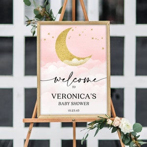 May include: A rectangular sign in a gold frame featuring a pink watercolor sky with a gold glitter crescent moon, clouds, and stars. The text reads: welcome TO VERONICA'S BABY SHOWER 01.23.45. The sign is displayed on a wooden easel.