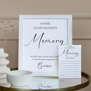 Puede incluir: Un letrero enmarcado blanco y una tarjeta más pequeña con el texto "Comparte tu recuerdo favorito" en escritura elegante. El letrero incluye el nombre "Brian". La tarjeta tiene líneas para escribir y las palabras "Mejores deseos".