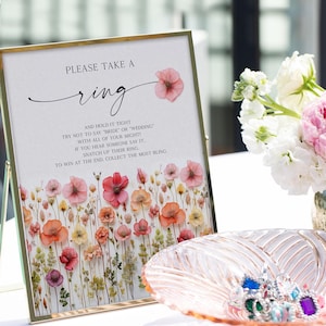 K&ouml;nnte beinhalten: Ein gerahmtes Schild mit dem Text "PLEASE TAKE A ring" und floralen Illustrationen. Eine dekorative Schale enth&auml;lt mehrere bunte Ringe. Das Schild hat einen goldenen Rahmen und steht auf einem Tisch. Der Text auf dem Schild lautet "AND HOLD IT TIGHT..."