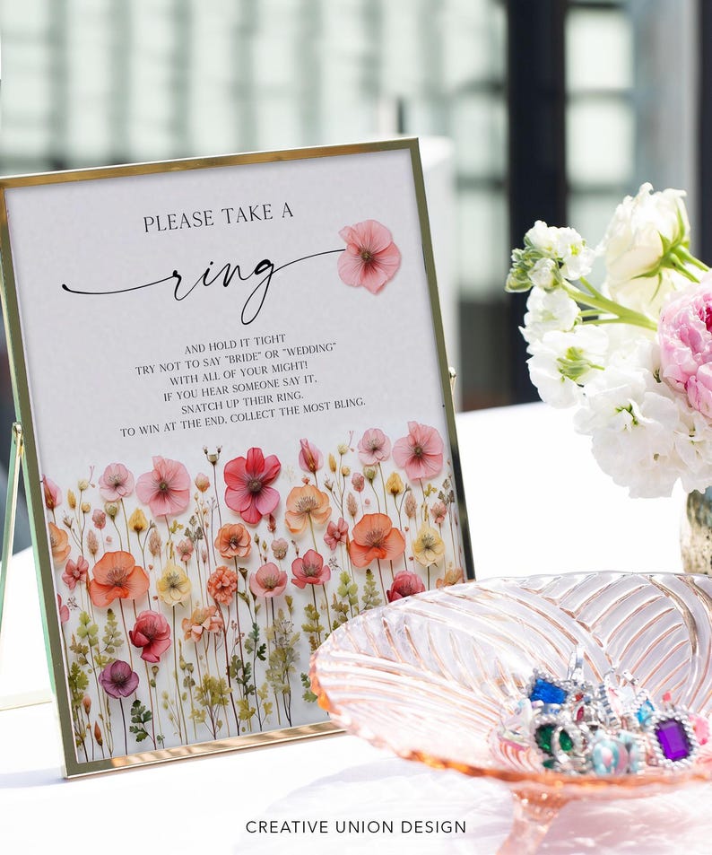 K&ouml;nnte beinhalten: Ein gerahmtes Schild mit dem Text "PLEASE TAKE A ring" und floralen Illustrationen. Eine dekorative Schale enth&auml;lt mehrere bunte Ringe. Das Schild hat einen goldenen Rahmen und steht auf einem Tisch. Der Text auf dem Schild lautet "AND HOLD IT TIGHT..."