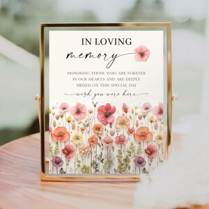May include: A framed memorial sign with the words "IN LOVING MEMORY" and "wish you were here." The sign features a floral design with pink, red, and orange flowers. The sign is in a gold frame and sits on a wooden surface.