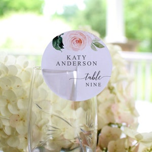 May include: A white round table number card with a pink watercolor floral design. The card says "Katy Anderson" and "table nine".