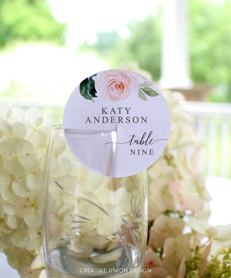 May include: A white round table number card with a pink watercolor floral design. The card says "Katy Anderson" and "table nine".