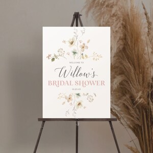 May include: A welcome sign for Willow's bridal shower, displayed on a wooden easel. The sign features floral watercolor illustrations in soft colors and the text "Welcome to Willow's Bridal Shower" in elegant script and a light pink font. The date 01.23.45 is also included.