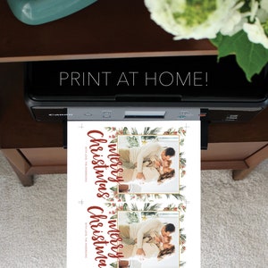 May include: A printer is shown printing two Christmas cards. The cards feature a family photo within a gold frame, surrounded by a festive border of greenery and berries. The words "Merry Christmas" are written in a flowing red script, with the text "Print at Home!" above the printer.