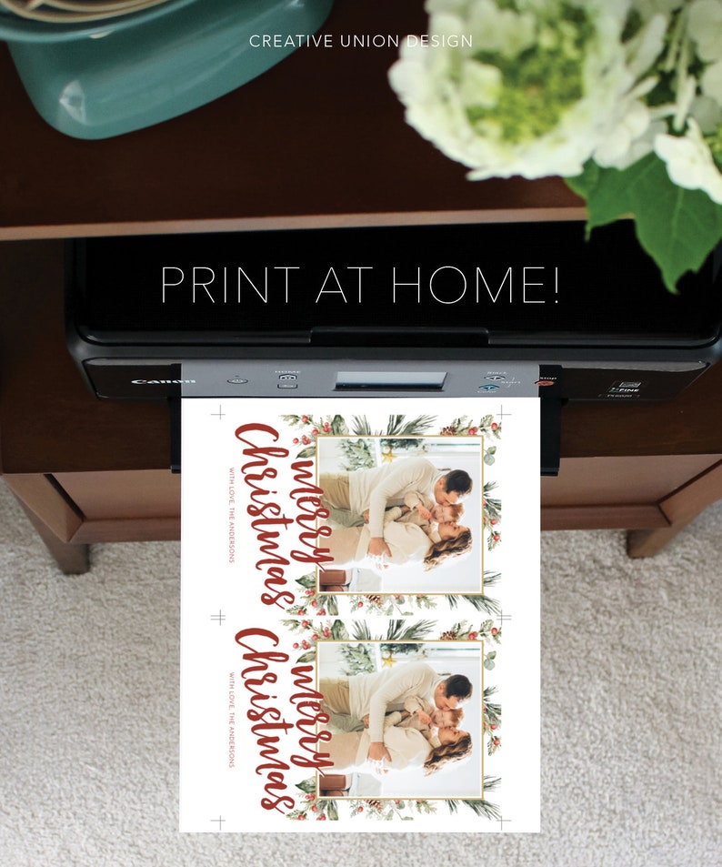 May include: A printer is shown printing two Christmas cards. The cards feature a family photo within a gold frame, surrounded by a festive border of greenery and berries. The words "Merry Christmas" are written in a flowing red script, with the text "Print at Home!" above the printer.