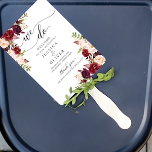 May include: A white wedding fan with floral accents in burgundy and blush tones. The fan features the words "we do", "Welcome to our beginning", and the names Jessica & Oliver. The date and location are also printed on the fan.