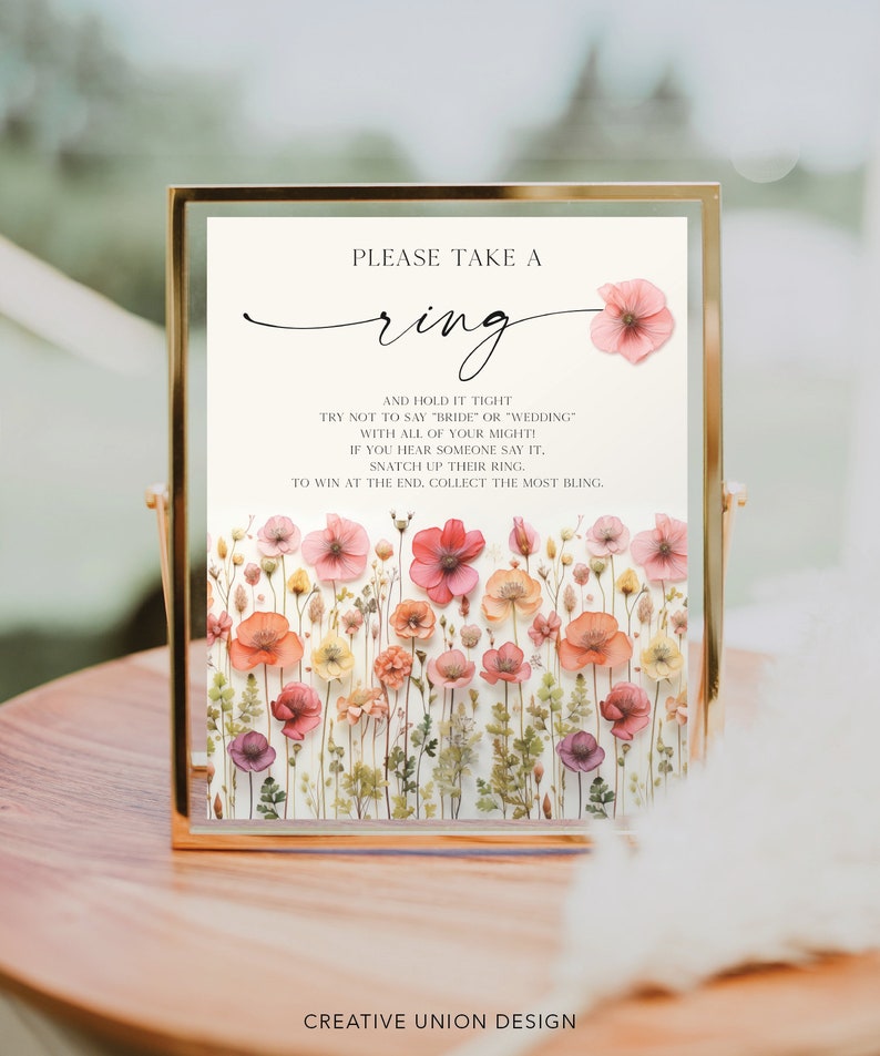 May include: A sign with a floral background and the text "Please take a ring and hold it tight. Try not to say "bride" or "wedding" with all of your might! If you hear someone say it, snatch up their ring. To win at the end, collect the most bling." The sign is in a gold frame.