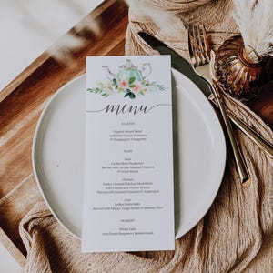 May include: A wedding menu card with a floral teapot design, placed on a white plate with silverware. The menu includes starter, main, and dessert options. The card is on a wooden surface with a beige cloth.