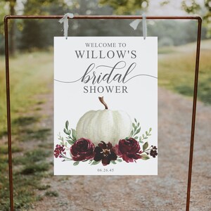 May include: White bridal shower sign with the text "Welcome to Willow's bridal shower" and a white pumpkin with burgundy flowers and greenery. The date "06.26.45" is printed at the bottom of the sign.
