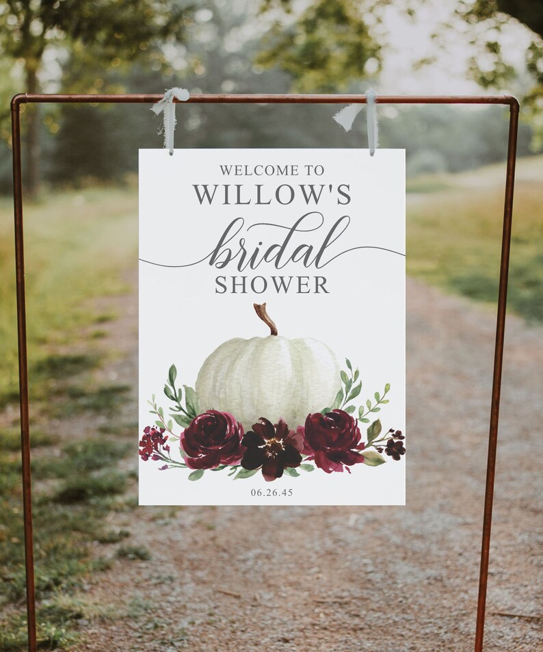 May include: White bridal shower sign with the text "Welcome to Willow's bridal shower" and a white pumpkin with burgundy flowers and greenery. The date "06.26.45" is printed at the bottom of the sign.