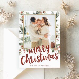 May include: A Christmas card with a family photo inside a gold-framed border of greenery and red berries. The words "Merry Christmas" are in red script, with "With love, The Andersons" below. A white envelope and silver ornaments are in the background.
