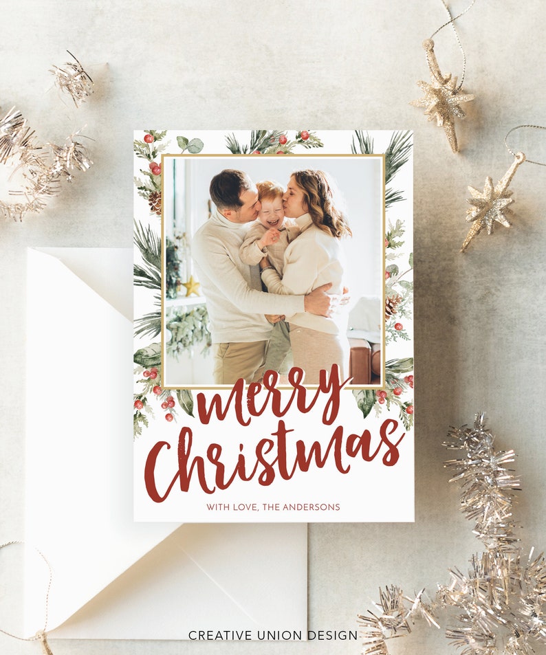 May include: A Christmas card with a family photo inside a gold-framed border of greenery and red berries. The words "Merry Christmas" are in red script, with "With love, The Andersons" below. A white envelope and silver ornaments are in the background.