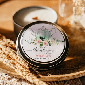May include: A small, silver tin candle with a white label. The label features a watercolor illustration of a teapot with pink and white flowers. The text on the label reads "thank you Willow's Bridal Shower".