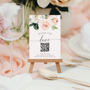 Può includere: Un piccolo cavalletto in legno sostiene un cartello con dettagli floreali e le parole "Share the Love". Sotto il testo c'è un codice QR. Il cartello è su un tavolo con un bicchiere e posate.