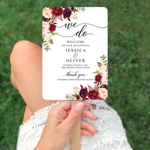 May include: A rectangular wedding fan with a wooden handle. The fan features the words "we do" in elegant script, along with the names Jessica & Oliver, and the date August 23, 2045. Floral accents in burgundy and blush pink adorn the edges.