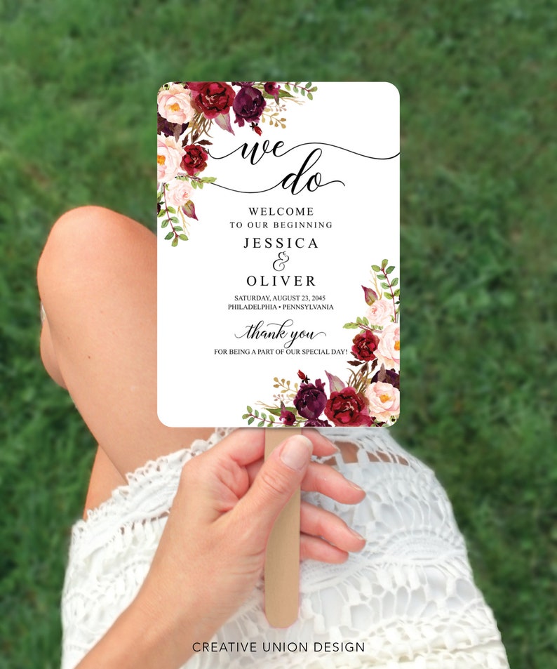 May include: A rectangular wedding fan with a wooden handle. The fan features the words "we do" in elegant script, along with the names Jessica & Oliver, and the date August 23, 2045. Floral accents in burgundy and blush pink adorn the edges.
