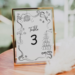 May include: A gold-framed table number card with black ink illustrations. The card features a disco ball, a vase with flowers, a champagne tower, and the text "Table 3".