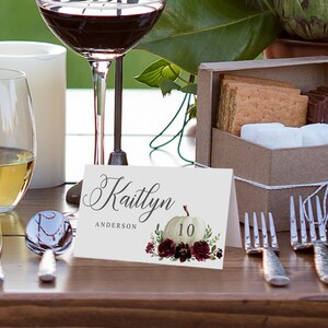 May include: A white place card with the name "Kaitlyn" and "Anderson" in elegant script. A watercolor pumpkin and floral design with the number 10 is featured. The card is set on a wooden table with wine glasses and silverware.