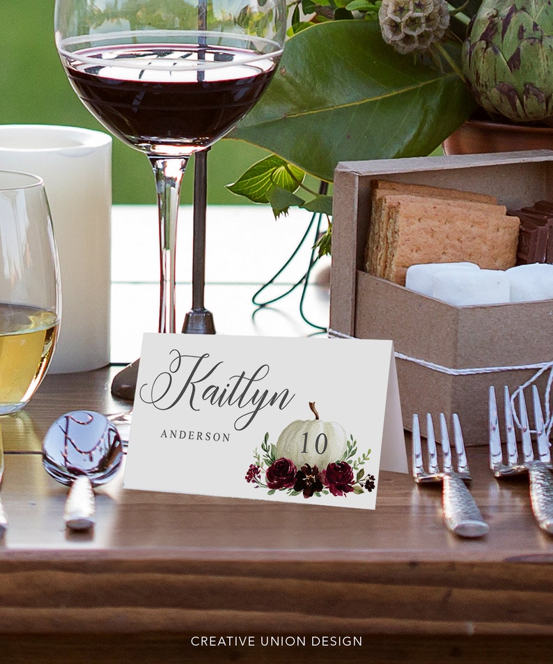 May include: A white place card with the name "Kaitlyn" and "Anderson" in elegant script. A watercolor pumpkin and floral design with the number 10 is featured. The card is set on a wooden table with wine glasses and silverware.