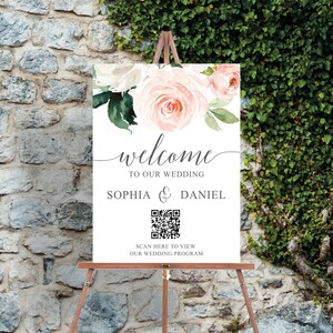 Op de afbeelding: Een wit welkomstschild voor een bruiloft met roze en witte bloemenillustraties. Het bord staat "welkom op onze bruiloft Sophia & Daniel" en bevat een QR-code met de tekst "Scan hier om ons huwelijksprogramma te bekijken".