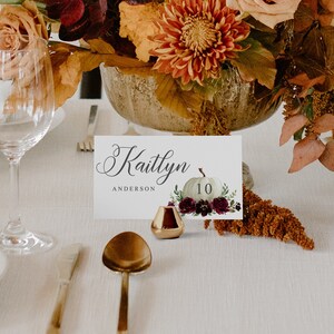 May include: A table setting with a floral centerpiece and place card. The place card features the name "Kaitlyn Anderson" and a pumpkin illustration with the number 10. A small gold candle holder and gold cutlery are also visible.