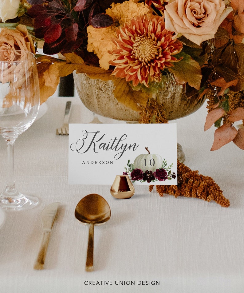 May include: A table setting with a floral centerpiece and place card. The place card features the name "Kaitlyn Anderson" and a pumpkin illustration with the number 10. A small gold candle holder and gold cutlery are also visible.