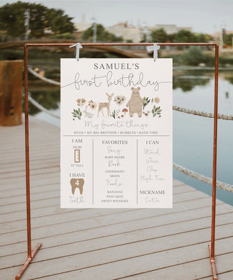 May include: A white printable first birthday sign with a woodland theme. The sign features a bear, deer, and rabbit surrounded by flowers and mushrooms. The sign includes the name Samuel, the age, and a list of the child's favorite things, including songs, books, foods, and activities.