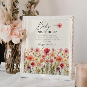 May include: A white framed sign with the text "Baby Sock Hunt" and "Baby socks have been hidden throughout the baby shower. Whoever finds the most at the end, wins a prize! Happy Hunting!" The sign has a floral design with pressed flowers in shades of pink, orange, and yellow.