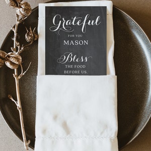 Puede incluir: Un cubierto con un plato gris oscuro, una servilleta de lino blanca y una tarjeta negra con escritura blanca que dice "Grateful for you Mason Bless the food before us". Un tallo de planta seca est&aacute; a la izquierda.