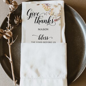 May include: A white place setting with a folded napkin and a card that reads "Give thanks, Mason, bless the food before us." The card has a floral design in the upper right corner. A decorative stem is on the left.