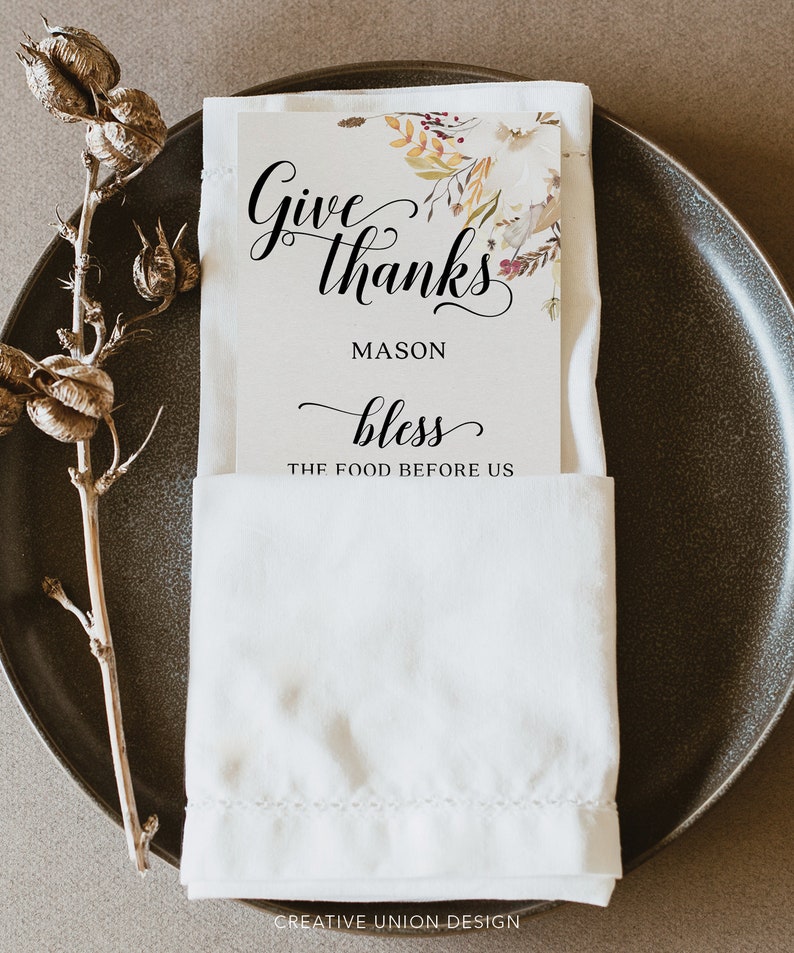 May include: A white place setting with a folded napkin and a card that reads "Give thanks, Mason, bless the food before us." The card has a floral design in the upper right corner. A decorative stem is on the left.