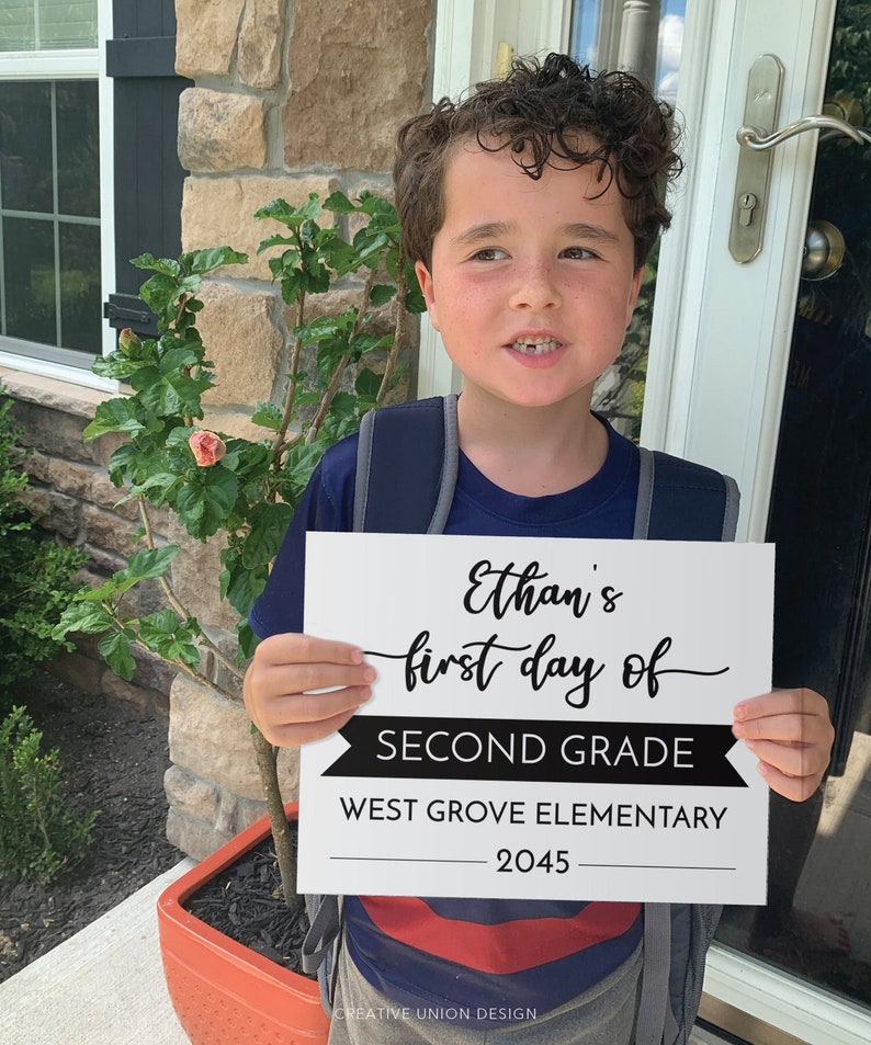 May include: A rectangular white sign with black text that reads Ethan's first day of SECOND GRADE WEST GROVE ELEMENTARY 2045. The sign features a black banner graphic behind the words SECOND GRADE and is held by a child wearing a navy blue shirt.