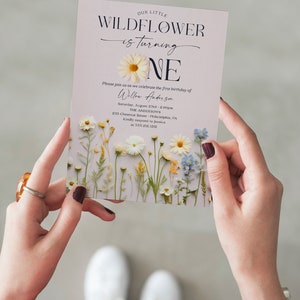 May include: A light purple invitation with a floral design featuring white daisies, yellow wildflowers, and blue flowers. The text reads "Our Little Wildflower is turning One" with a large white daisy and the number "One" in black text. The invitation includes the name "Willow Anderson", the date "Saturday, August 23rd - 2:00pm", the location "The Anderson's 235 Chestnut Street Philadelphia, PA", and the contact information "Kindly respond to Jessica at 535.212.1212". The bottom of the invitation reads "Creative Union Design".